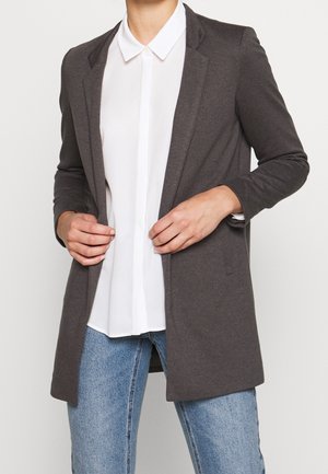 Person trägt ein dunkelgraues offenes Sakko über einem weißen Hemd mit Kragen und hellblauen Jeans und richtet das Sakko mit beiden Händen.