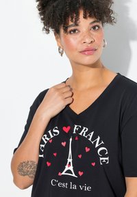 Černé tričko s véčkovým výstřihem, na kterém je bílý text "PARIS ♥ FRANCE" s motivem červeného srdce a grafikou Eiffelovy věže. Měkká, pohodlná látka.