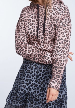 Vrouw met een roze hoodie met luipaardprint, gecombineerd met een blauwe rok met luipaardprint, armen gekruist, tegen een eenvoudige lichte achtergrond.