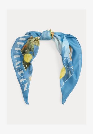 Pañuelo de seda azul con estampados de limones y hojas, anudado en el centro, con texto grande blanco parcialmente visible en el borde.
