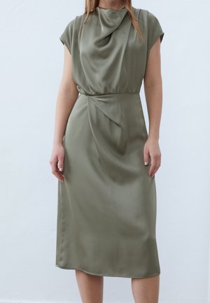 Kobieta w oliwkowo-zielonej satynowej sukience midi z marszczonym dekoltem i detalem zebranym w talii, stojąca na jasnym, jednolitym tle.