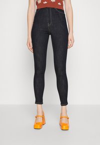 Jeans de cintura alta em tom escuro, com corte slim e costura em contraste, combinados com sapatos plataforma laranja brilhantes. Blusa simples com padrão floral.