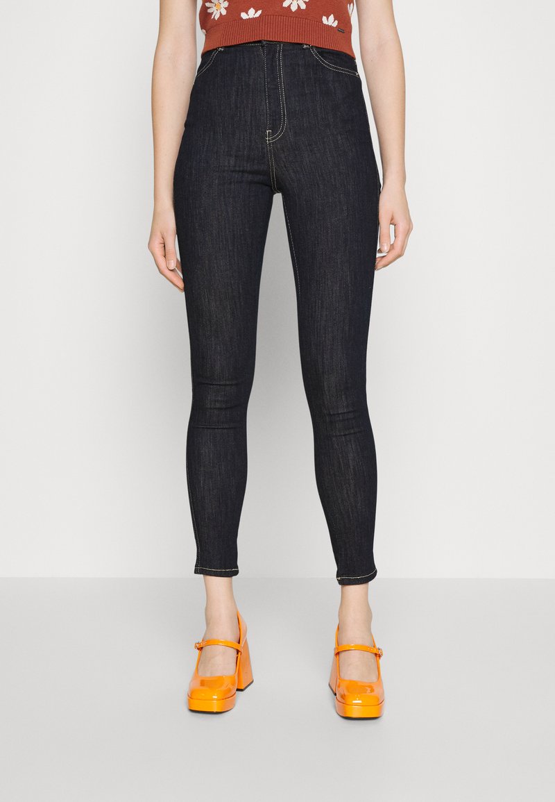 Jeans de cintura alta em tom escuro, com corte slim e costura em contraste, combinados com sapatos plataforma laranja brilhantes. Blusa simples com padrão floral.