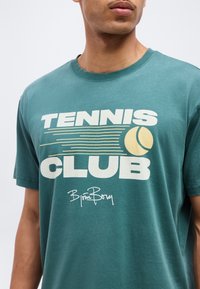 Άνδρας που φοράει πετρατζέν μπλουζάκι με το κείμενο "TENNIS CLUB", γραφικό με κίτρινη μπάλα τένις και υπογραφή του Björn Borg στο μπροστινό μέρος.