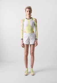 Strak aansluitende bodysuit in wit en neon geel met uitgesneden schouders, gecombineerd met witte shorts en groene veterschoenen.