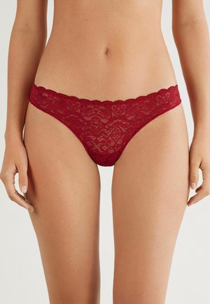String en dentelle rouge avec des bords festonnés et des motifs floraux complexes. La texture transparente met en valeur les détails de conception dans une coupe taille basse.