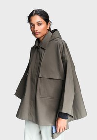 Femme aux cheveux foncés porte une cape à capuche oversize de couleur olive avec de grandes poches sur un pantalon clair.