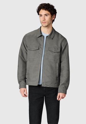 Junger Mann mit lockigem Haar, der eine graue Freizeitjacke über einem hellblauen Hemd und schwarzen Hosen trägt, steht vor einem schlichten Hintergrund.