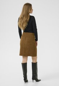 Kvinde med langt blondt hår iført en sort langærmet top, brun knælang nederdel med baglommer og sorte knæhøje hæle støvler, vendt sidelæns.