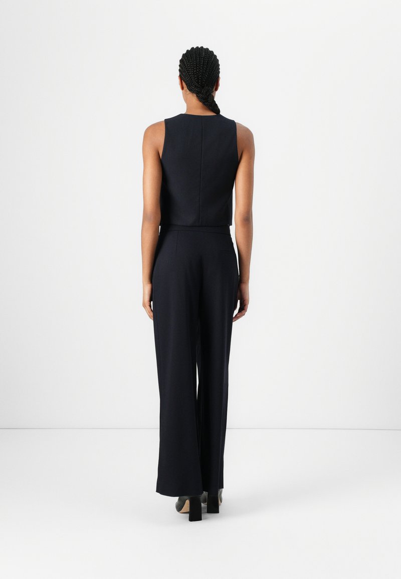 Marineblauwe aansluitende jumpsuit met een mouwloos ontwerp, wijde benen, een soepele textuur en een hoge nek. De achteraanzicht toont de naaddetails en zijzakken.
