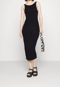 Černé, bezrukávové midi šaty s těsným střihem. Modelka má na sobě masivní černé sandály a drží vzorovanou tašku s monochromatickým designem.