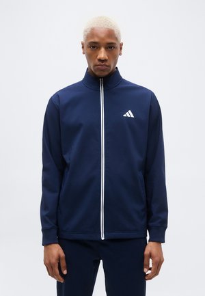 Mann mit kurzem blondem Haar, der eine marineblaue Jacke mit Reißverschluss und weißem Logo trägt, vor einem einfarbigen hellen Hintergrund steht.