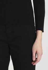 Pull noir à côtes avec des poignets ajustés et un ourlet droit, associé à un pantalon noir. La texture est lisse et légèrement extensible.
