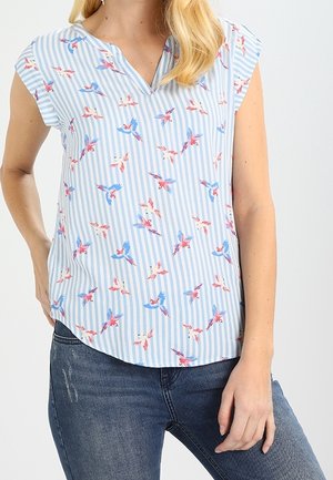Woman wearing a light blue and white vertical striped blouse with red and blue bird prints, paired with dark blue jeans.