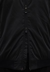 Gros plan sur une veste noire avec une double fermeture éclair, une fermeture partiellement ouverte et l'autre complètement fermée, montrant la texture du tissu.