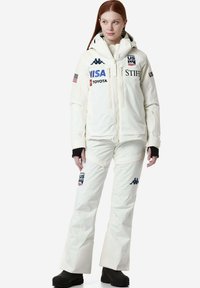 Giacca e pantaloni da sci bianchi con logo, chiusure a zip e tasche. La giacca ha un cappuccio e i pantaloni hanno una linea dritta. Guanti e stivali neri.