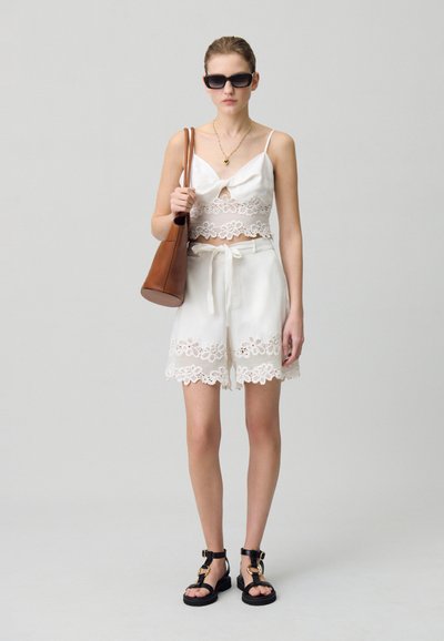 Top corto in pizzo bianco con un fiocco, abbinato a pantaloncini bianchi con bordo in pizzo. La modella porta una borsa marrone e indossa sandali neri.