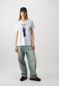 Witte katoenen t-shirt met grafische print en tekst, gecombineerd met lichtblauwe baggy jeans. Zwarte pet en sneakers met rode accenten.