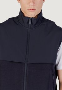 Gilet senza maniche blu navy con colletto alto, chiusura con zip e pannelli di tessuto a contrasto. Presenta una tasca laterale per utility. Texture liscia.