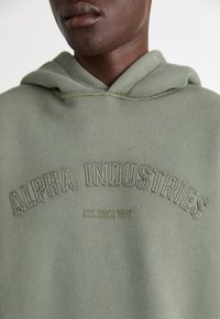 Sweat à capuche vert clair avec broderie "ALPHA INDUSTRIES" et "EST. SINCE 1959" en fil vert foncé. Texture de tissu douce.