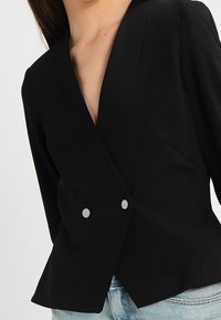 Svart kavaj med dubbla silverknappar som bärs över ljusblå denimjeans på en person med långt mörkt hår.