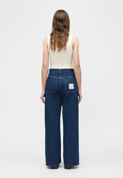 Γυναίκα με μακριά καστανά μαλλιά, που φοράει λευκό αμάνικο τοπ και ψηλόμεσα, φαρδιά, σκούρα μπλε τζιν παντελόνια, που στέκεται γυρισμένη με την πλάτη.