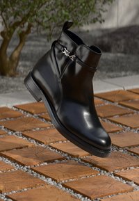 Schwarze Leder-Ankle-Boots mit seitlichem Riemen und silbernem Logo-Detail, präsentiert über einem Holzblockweg mit Kies und verschwommenem grünen Hintergrund.