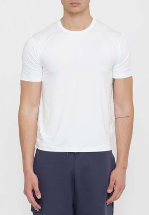 Vista frontale di una persona che indossa una t-shirt bianca semplice a maniche corte e pantaloni blu scuro, con tatuaggi visibili sugli avambracci.