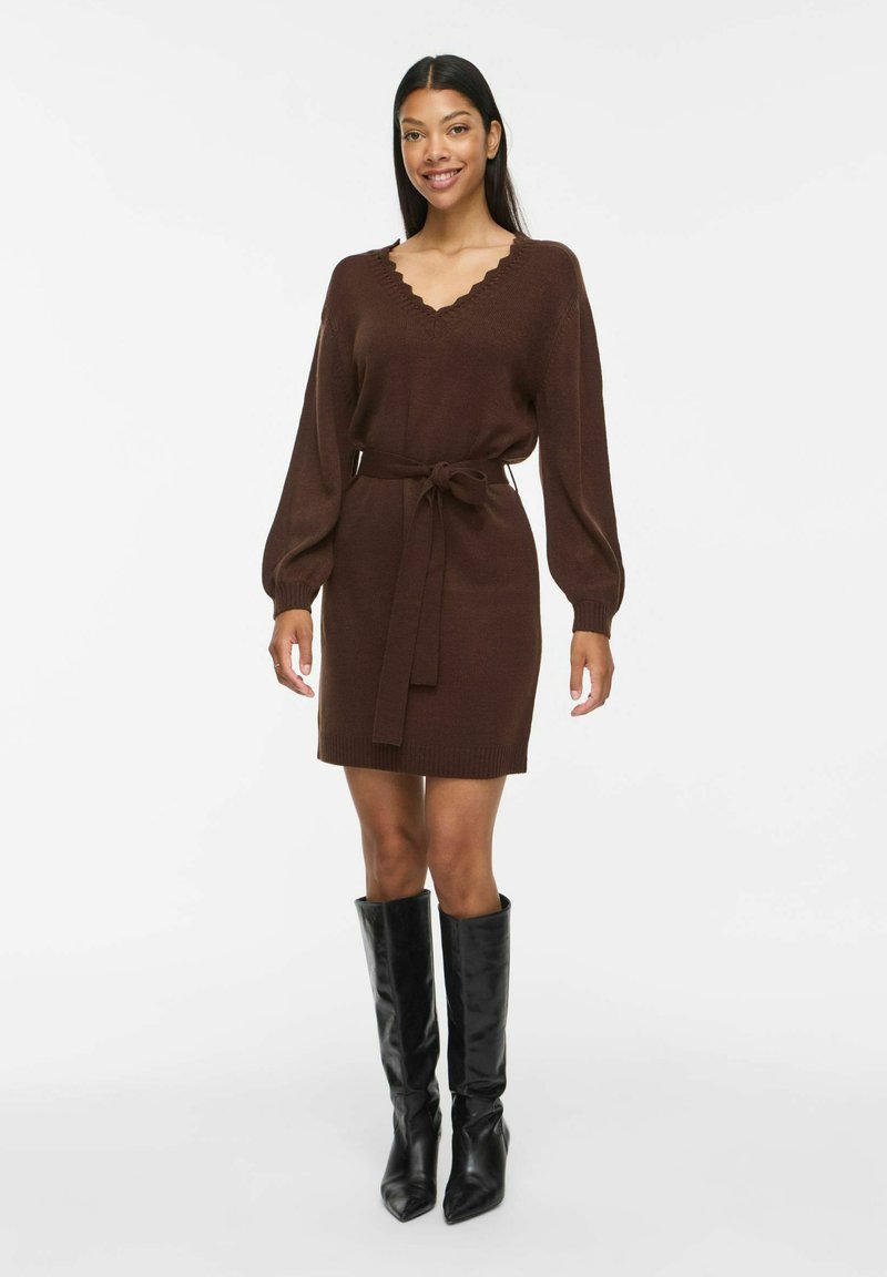 Femme portant une robe en maille marron à manches longues avec un col en V et une ceinture, associée à des bottes noires hautes à bout pointu, debout sur un fond blanc.