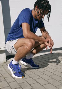 Blaue Sport Schuhe mit strukturiertem Stoff, gelben Akzenten und weißen Sohlen. Getragen mit grauen Shorts und einem passenden blauen T-Shirt auf Asphalt.