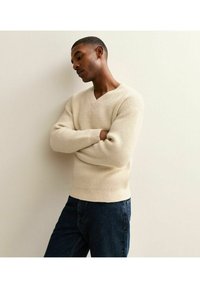 Gestrickter cremefarbener Pullover mit V-Ausschnitt und gerippter Textur, kombiniert mit dunklen Jeans. Hat lange Ärmel und eine taillierte Silhouette.