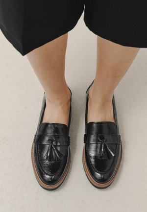 Pieds portant des mocassins en cuir noir brillant avec des glands et des semelles marron, debout sur un sol clair.