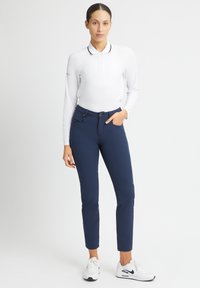 White long-sleeve polo shirt with a collar and black trim, paired with navy slim-fit pants. Model wears white sneakers. Simple design, no patterns.