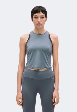 Jonge vrouw met kort donker haar die een mouwloos grijs sporttopje en bijpassende legging draagt tegen een effen achtergrond.