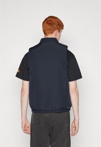 Ναυτικό μπλε ελαφρύ γιλέκο με υφασματική υφή, όρθιο γιακά και ελαστική μανσέτα. Φοριέται πάνω από ένα μαύρο πουκάμισο, που διαθέτει λογότυπο στο μανίκι.