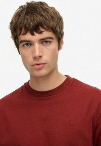 Ung mand med kort brunt hår og blå øjne, iført en ensfarvet mørkerød t-shirt med rund hals, neutral ansigtsudtryk, hvid baggrund.