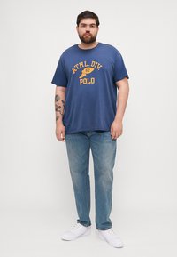 Tricou din bumbac bleu marin, cu un logo imprimat galben care citește "ATHL.DIV.67 POLO", asortat cu blugi albaștri deschiși și adidași albi.