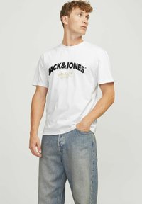 Camiseta de algodón blanca con un audaz logo negro "JACK&JONES" y texto con acentos dorados. El modelo la combina con pantalones vaqueros de denim azul.