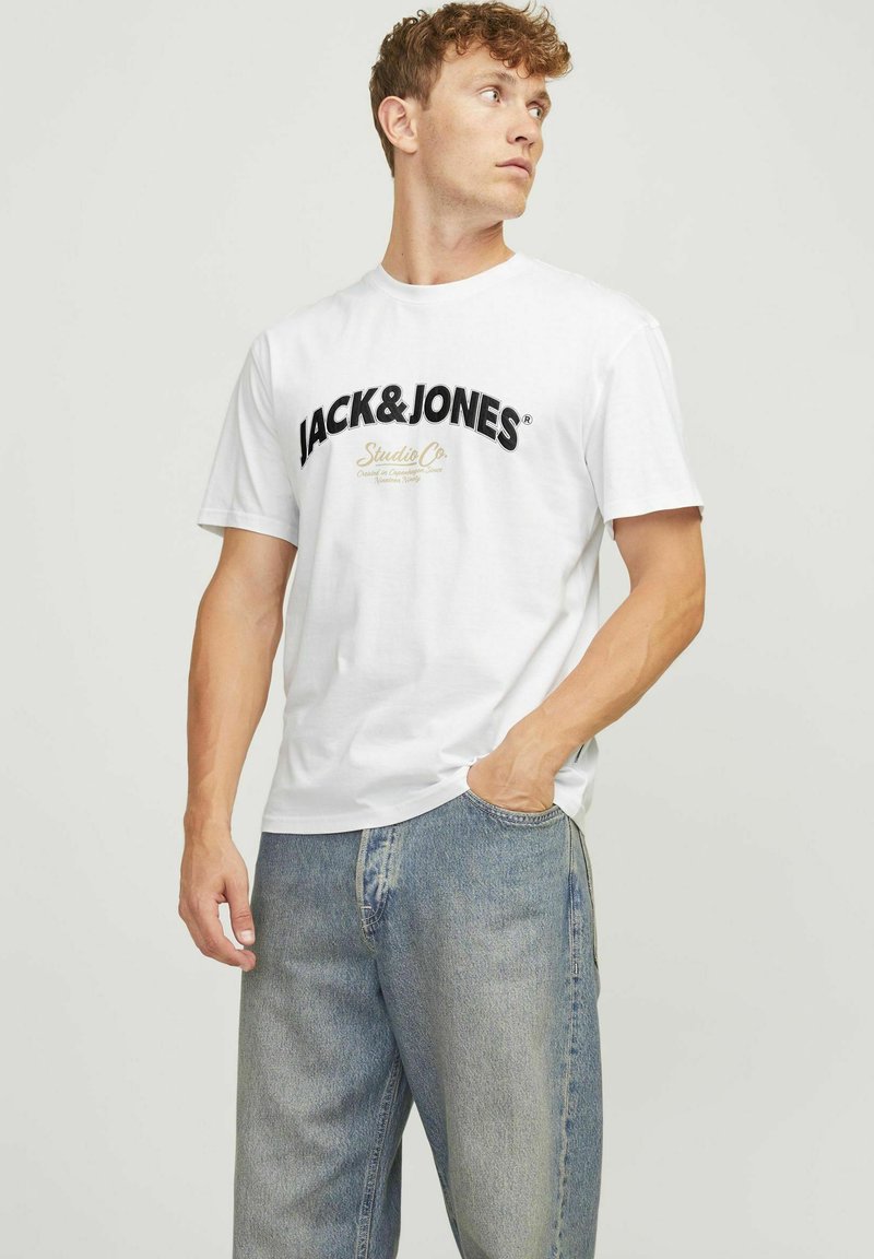 Camiseta de algodón blanca con un audaz logo negro "JACK&JONES" y texto con acentos dorados. El modelo la combina con pantalones vaqueros de denim azul.
