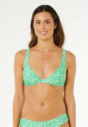 Jonge vrouw met groene bloemenbikini, glimlachend, staand tegen een witte achtergrond.