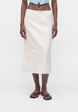 Femme portant une jupe midi blanche à rayures horizontales avec une taille à cordon, associée à des sandales marron et un top court bleu clair.