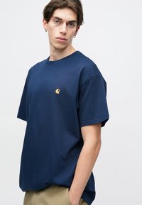 Mornarsko modra bombažna majica s kratkimi rokavi, okroglim ovratnikom in majhnim rumenim logotipom na prsih. Gladka tekstura in sproščen kroj.