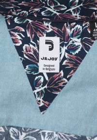 Bloemmotief stof in marineblauw met roze accenten, gecombineerd met een geweven lichtblauwe stof. Labels met de tekst "J&JOY" en "Ontworpen in België."