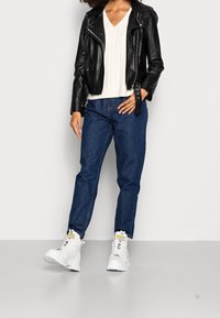Schwarze Leder-Bikerjacke, cremefarbener Grobstrickpullover, blaue high-waisted Jeans und klobige weiße Sneakers mit gelben Akzenten.