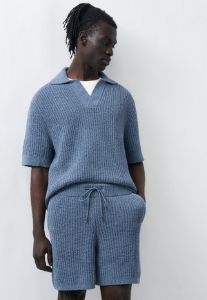 Sötét bőrű férfi dreadlockkal, kék kötött, rövid ujjú galléros felsőt és hozzáillő, zsinóros rövidnadrágot visel, és egy sima fal előtt áll.