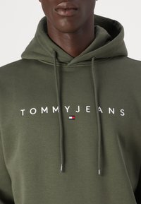 Olive spalvos džemperis su kapuce ir raišteliais, su išsiuvinėtu "TOMMYJEANS" balta spalva ir maža raudona, balta bei mėlyna logotipo etikete.