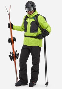 Veste de ski verte vif avec des accents noirs, pantalons noirs, bâtons de ski et skis avec des rayures orange. Comprend des gants et un casque.