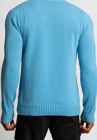 Man som bär en ljusblå stickad tröja med ribbade manschetter och nederkant, sedd bakifrån, i kombination med svarta byxor.