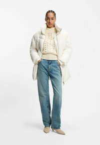 Uppblåst vit jacka med hög krage, krämfärgad stickad tröja med spetsmönster, ljusblå jeans och beige spetsiga klackskor.