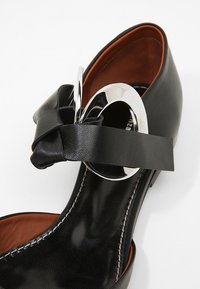 Zapato de cuero negro con un acento plateado redondeado, que presenta una correa ancha y un detalle de nudo en la parte frontal. Interior y costuras en marrón.